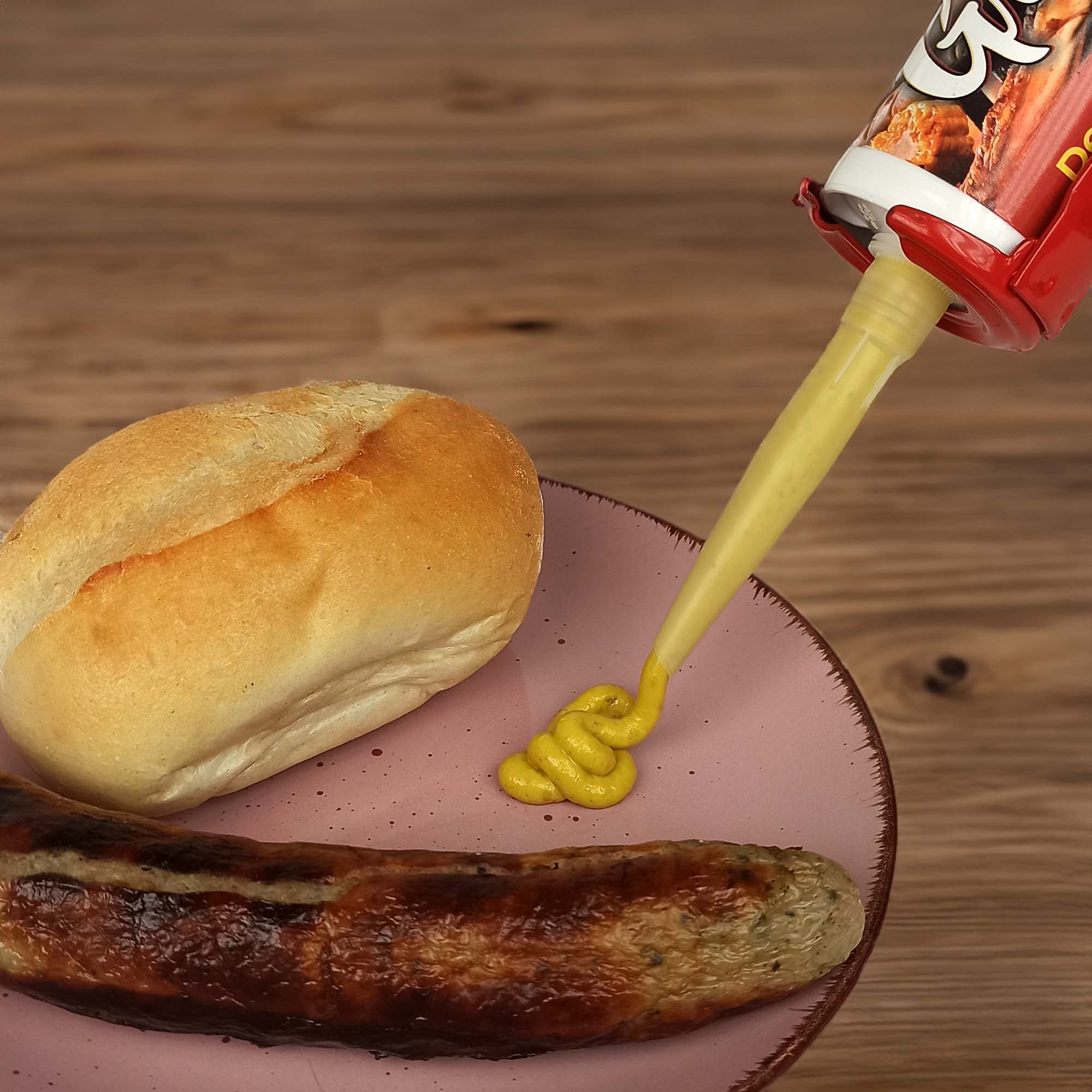 Roter Senfspender mit Quetschbeutel dosiert gelben Senf auf einen rosa Teller neben Brötchen und gegrillter Bratwurst